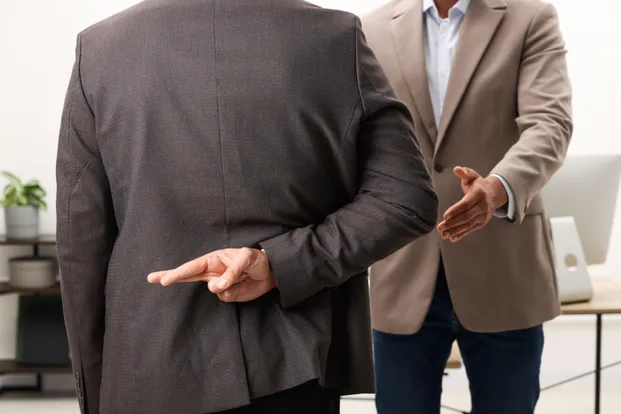 Two men in business attire stand facing each other, one gesturing with crossed fingers behind his back.